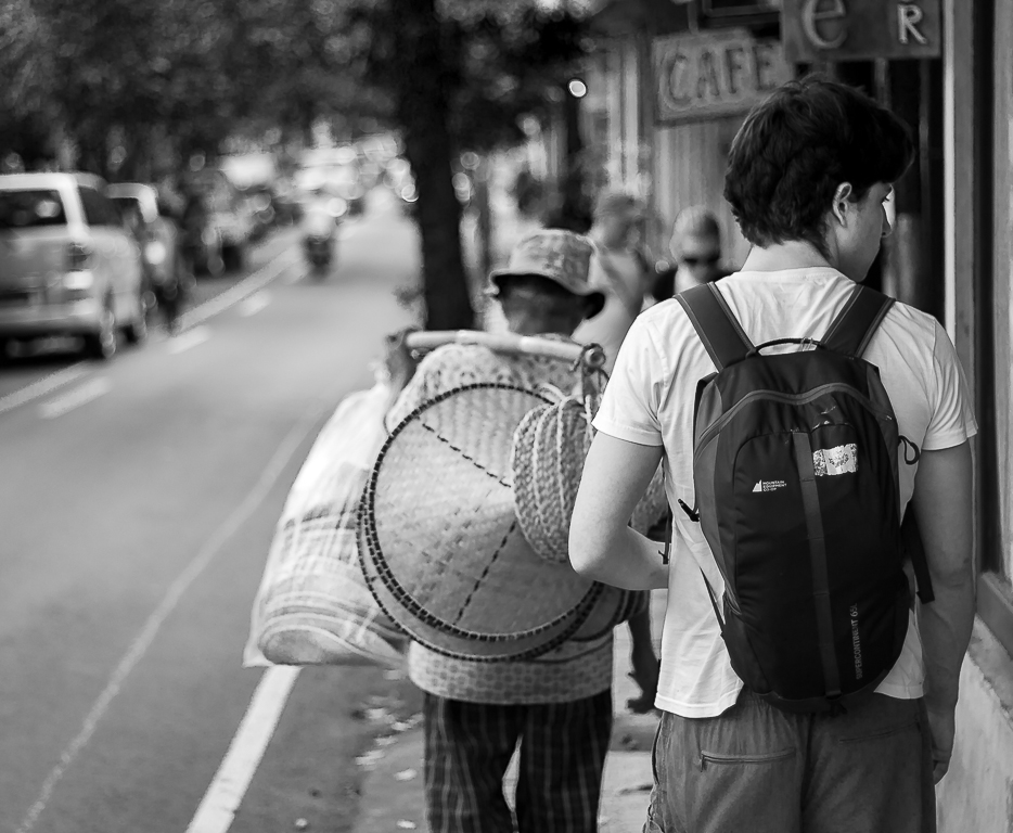 A porter with woven baskets passes a café, Bali